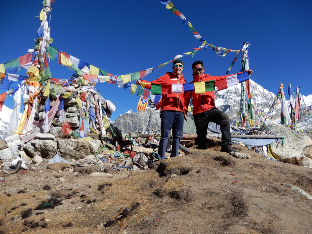 Zwei Bergsteiger von HÖHENBERGSTEIGEN zwischen farbigen Gebetsfahnen vor dem Himalaya
